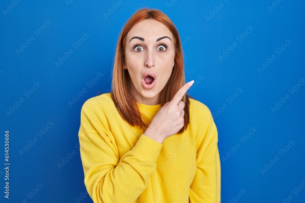 Young woman standing over blue background surprised pointing with finger to the side, open mouth amazed expression. Young woman standing over blue background surprised pointing with finger to the side, open mouth amazed expression.