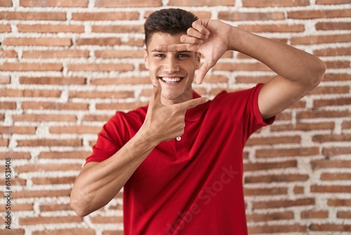 Young hispanic man standing over bricks wall smiling making frame with hands and fingers with happy face. creativity and photography concept.