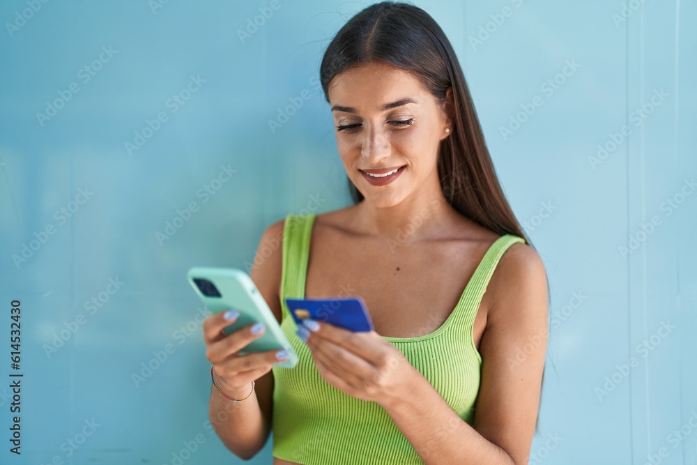 Young beautiful hispanic woman using smartphone and credit card over isolated blue background Young beautiful hispanic woman using smartphone and credit card over isolated blue background