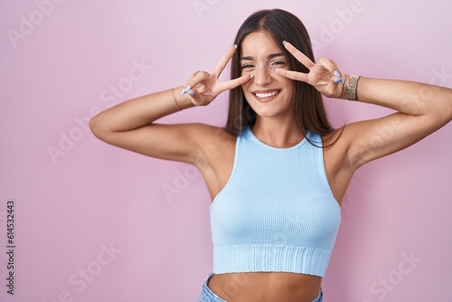 Young brunette woman standing over pink background doing peace symbol with fingers over face, smiling cheerful showing victory © Krakenimages.com
