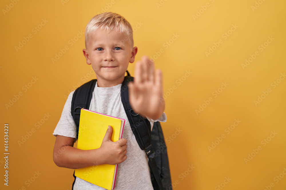 Little caucasian boy wearing student backpack and holding book with open hand doing stop sign with serious and confident expression, defense gesture Little caucasian boy wearing student backpack and holding book with open hand doing stop sign with serious and confident expression, defense gesture