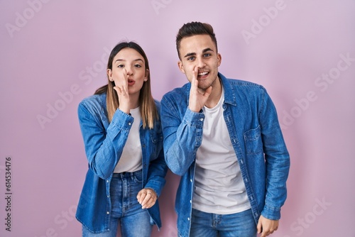 Young hispanic couple standing over pink background hand on mouth telling secret rumor, whispering malicious talk conversation