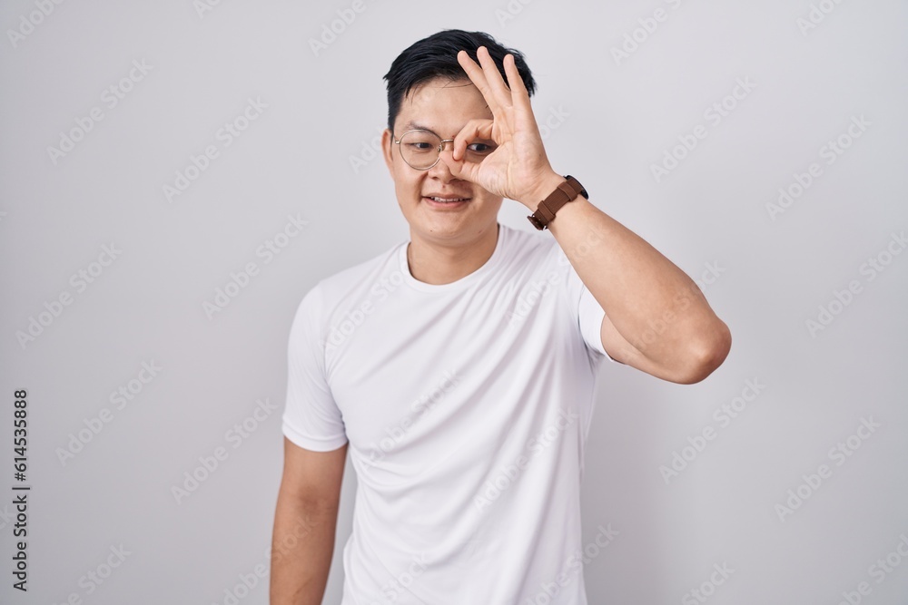 Young asian man standing over white background doing ok gesture with hand smiling, eye looking through fingers with happy face.