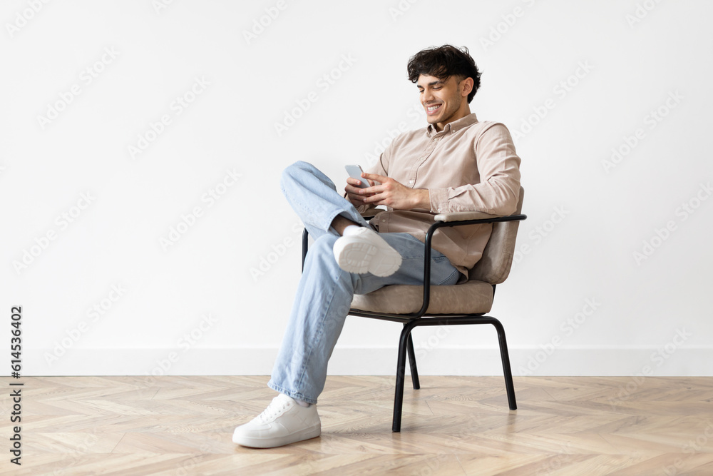 Happy Arab Guy Using Mobile Phone Sitting Over White Wall