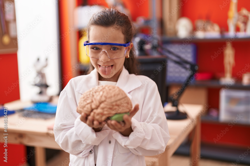 Fototapeta premium Little hispanic girl holding brain at science class at school sticking tongue out happy with funny expression.