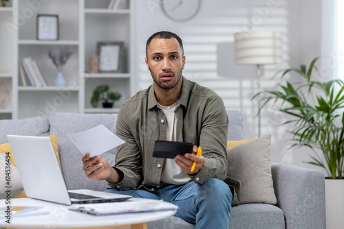 Portrait of a worried African-American man sitting at home on the couch with a laptop and documents. Calculates the family budget, financial income, loan and mortgage expenses
