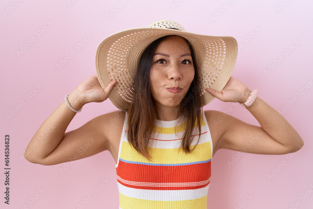 Middle age chinese woman wearing summer hat over pink background puffing cheeks with funny face. mouth inflated with air, catching air. Middle age chinese woman wearing summer hat over pink background puffing cheeks with funny face. mouth inflated with air, catching air.