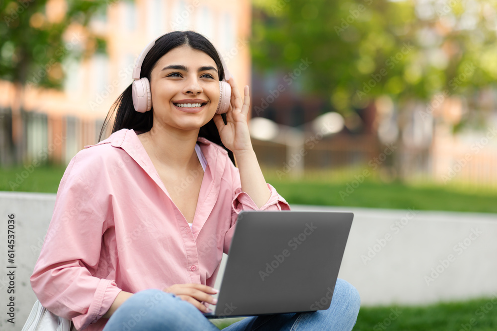 E-learning concept. Happy hispanic teen lady using laptop, wearing headphones listening to music, sitting in campus park E-learning concept. Happy hispanic teen lady using laptop, wearing headphones listening to music, sitting in campus park