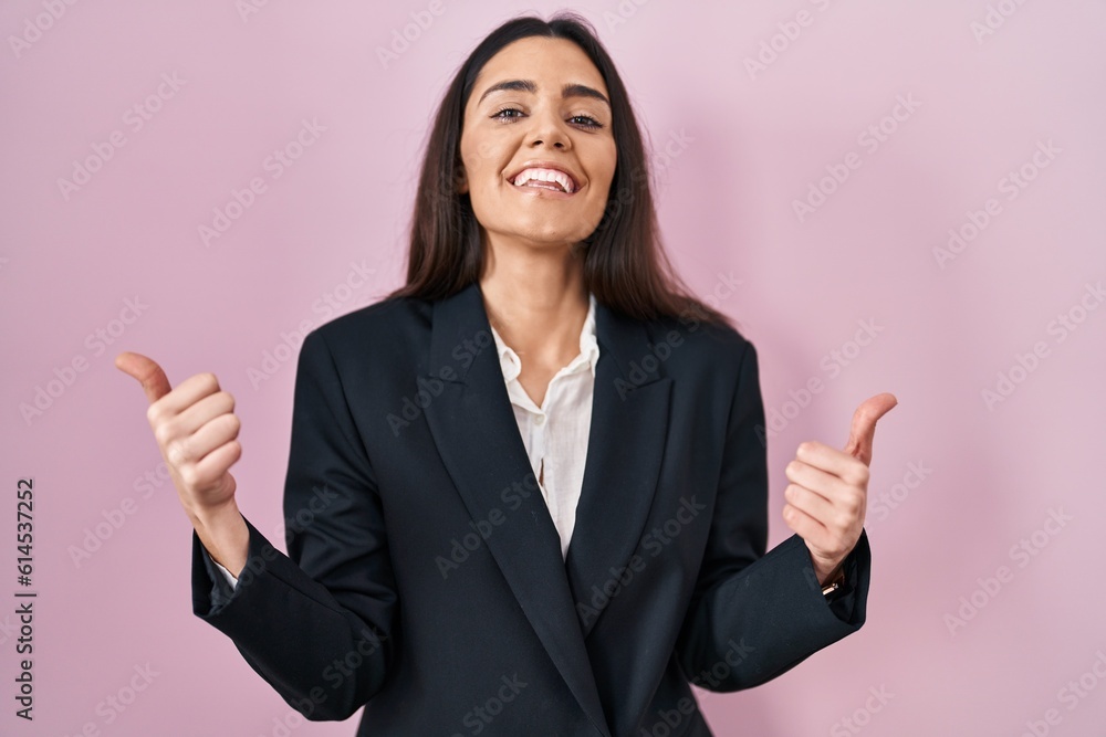 Young brunette woman wearing business style over pink background success sign doing positive gesture with hand, thumbs up smiling and happy. cheerful expression and winner gesture. Young brunette woman wearing business style over pink background success sign doing positive gesture with hand, thumbs up smiling and happy. cheerful expression and winner gesture.