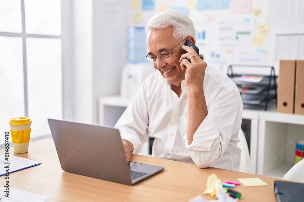 Middle age grey-haired man business worker using laptop talking on smartphone at office Middle age grey-haired man business worker using laptop talking on smartphone at office