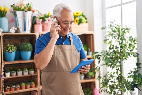 Middle age grey-haired man florist talking on smartphone using touchpad at florist