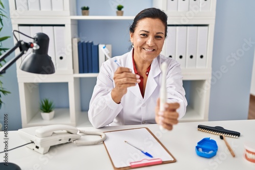 Middle age hispanic woman dentist holding toothbrushes at clinic