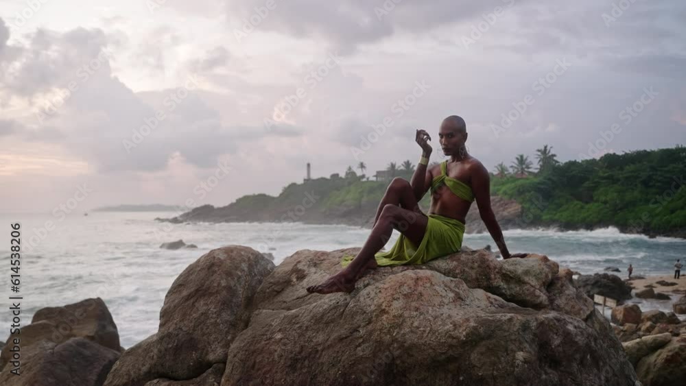 Non-binary black person poses gracefully sitting on top of rock in ocean at sunset slow motion. Trans ethnic fashion model in open tropical dress on peak by storm at dusk against sunrise Non-binary black person poses gracefully sitting on top of rock in ocean at sunset slow motion. Trans ethnic fashion model in open tropical dress on peak by storm at dusk against sunrise