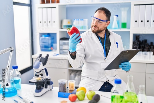 Young hispanic man scientist write on report working holding red pepper at laboratory
