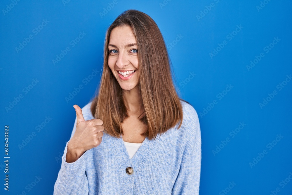 Fototapeta premium Young woman standing over blue background doing happy thumbs up gesture with hand. approving expression looking at the camera showing success.
