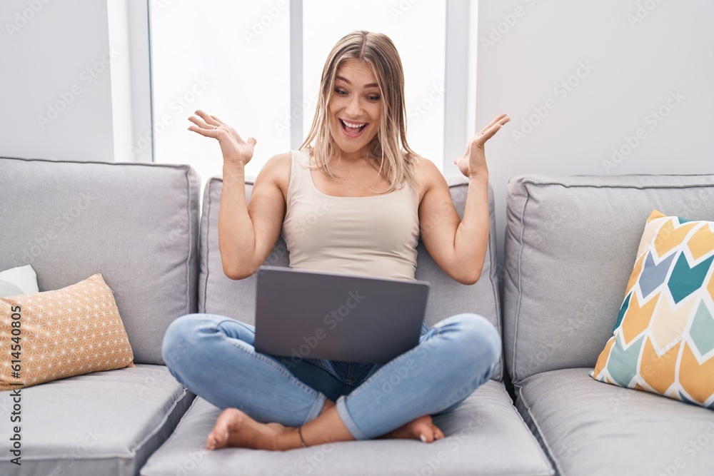 Blonde caucasian woman using laptop at home sitting on the sofa celebrating victory with happy smile and winner expression with raised hands Blonde caucasian woman using laptop at home sitting on the sofa celebrating victory with happy smile and winner expression with raised hands