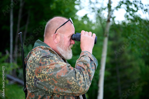 Gray-bearded senior hunter holding binoculars in hands and a rifle over his shoulder during hunting, walks through the forest, looks through binoculars and observes nature. Concept of hunt and travel photo