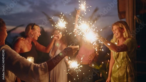 Diverse women dance and laugh with sparklers at seaside tropical hen party at sunset. Young smiling multiracial females have fun at Independence Day celebration by the sea with sparkling lights