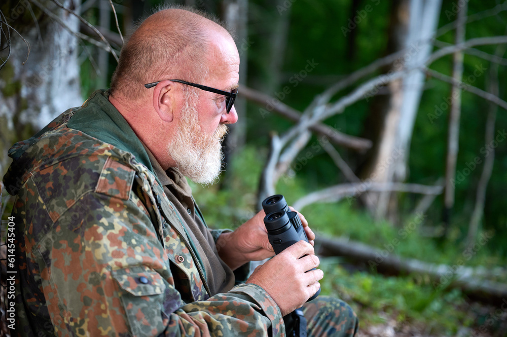 Gray-bearded senior hunter holding binoculars in hands and a rifle over his shoulder during hunting, walks through the forest, looks through binoculars and observes nature. Concept of hunt and travel