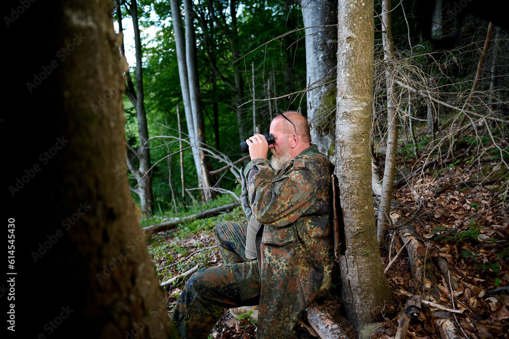 Gray-bearded senior hunter holding binoculars in hands and a rifle over his shoulder during hunting, walks through the forest, looks through binoculars and observes nature. Concept of hunt and travel