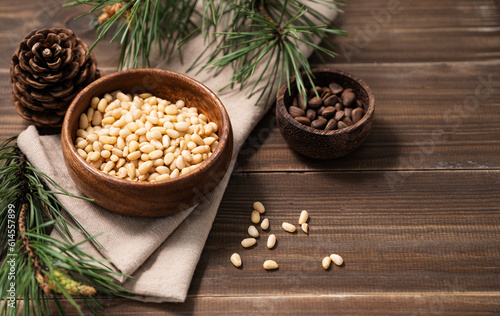 Pine nuts in a bowl on napkin and a handful of unpeeled nuts on a brown wooden background with a branch of pine needles. Healthy diet snack.