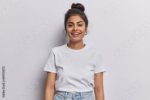 Horizontal shot of Indian woman with dark hair gathered in bun smiles pleasantly being in good mood dressed in casual t shirt and jeans isolated over white background. People and emotions concept photo