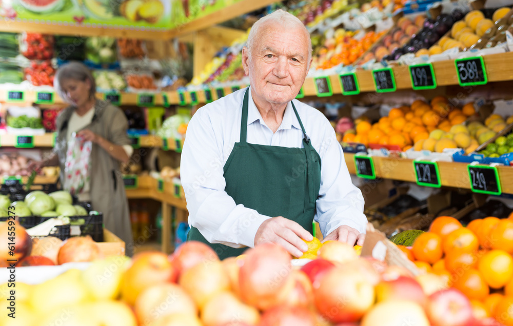 Old man greengrocer worker in apron standing in salesroom and setting out goods. Lady shopping in background..