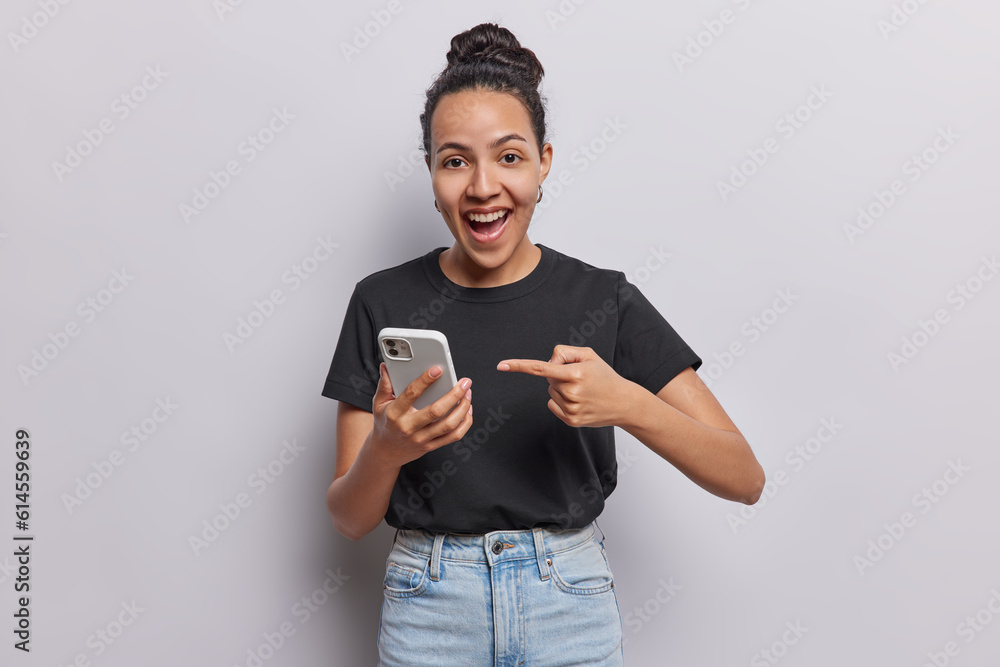 Young Latin positive woman with hair bun points at her smartphone keeps mouth opened laughs positively wears casual black t shirt and jeans poses against white background. Look at my new cellphone Young Latin positive woman with hair bun points at her smartphone keeps mouth opened laughs positively wears casual black t shirt and jeans poses against white background. Look at my new cellphone