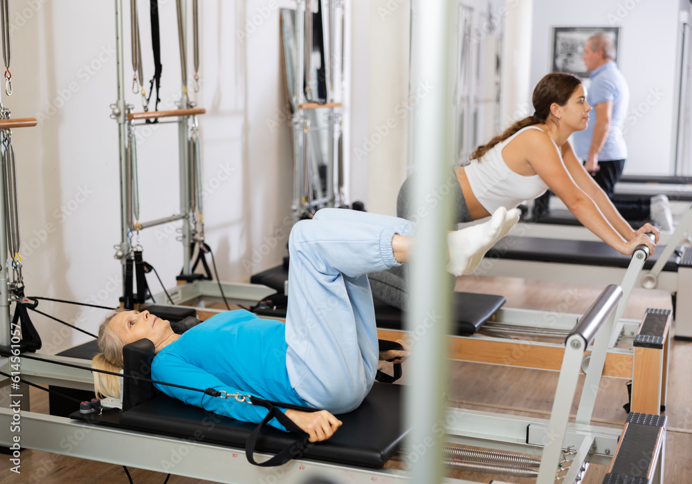 Positive aged woman doing Pilates exercises for lower back while lying on reformer bed in rehabilitation center. Fitness and wellness concept