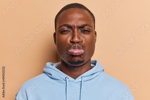 Studio shot of dark skinned man pouts lips frowns face expresses dissatisfaction being offended on someone wears blue sweatshirt isolated over brown background. Negative human emotions concept