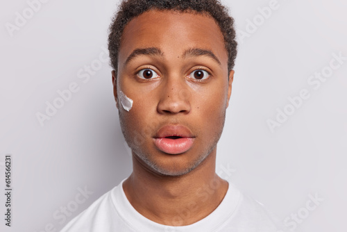 Close up shot of handsome middle aged dark skinned man applies face cream with care maintains healthy and nourished complexion keeps mouth opened stares impressed isolated over white background