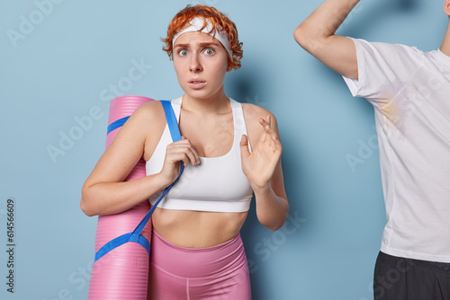 Shocked worried redhead sporty woman poses with rolled karemat stares impressed finishes her training poses in gym near unrecognizable sweaty man who shows biceps isolated over blue background