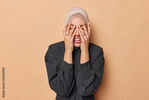 Horizontal shot of positive Muslim woman covers face with hands keeps eyes closed tries to hide herself wears traditional hijab and black jacket isolated over brown background. Islamic female model