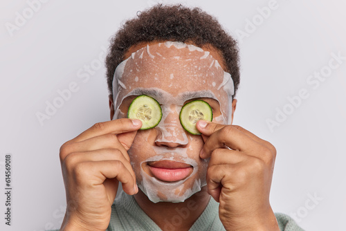 Self care and grooming concept. Curly haired unrecognizable man applies moisturising sheet mask covers eyes with cucumber slices for refreshing undergoes beauty procedures poses over white background