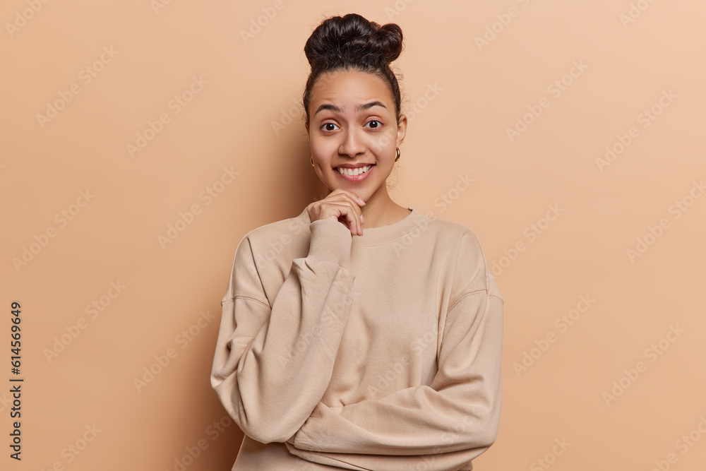 Fototapeta premium Waist up shot of cheerful Latin woman keeps hand on chin smiles gladfully has curious expression dressed in casual pullover isolated over brown background. Optimistic millennial female model