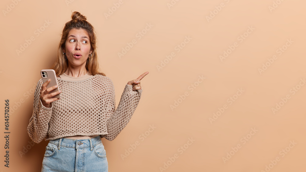 Young lady filled with excitement points her finger towards side inviting attention to free copy space has surprised expression holds mobile phone dressed in knitted jumper and jeans isolated Young lady filled with excitement points her finger towards side inviting attention to free copy space has surprised expression holds mobile phone dressed in knitted jumper and jeans isolated