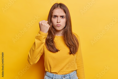 Horizontal shot of angry woman commands attention clenches her fist with fierce expression ready to assert authority and seek justice has irritated expression wears casual jumper and jeans isolated