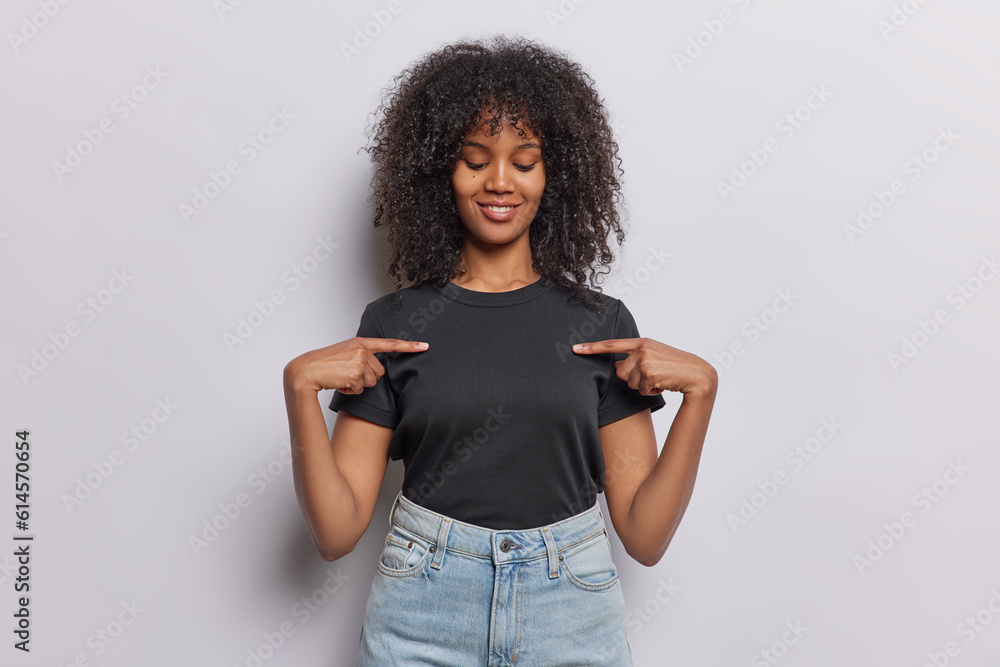 Φωτογραφία Studio shot of pleased curly haired young woman points at blank space of casual t shirt shows place for your logo or design isolated over white background