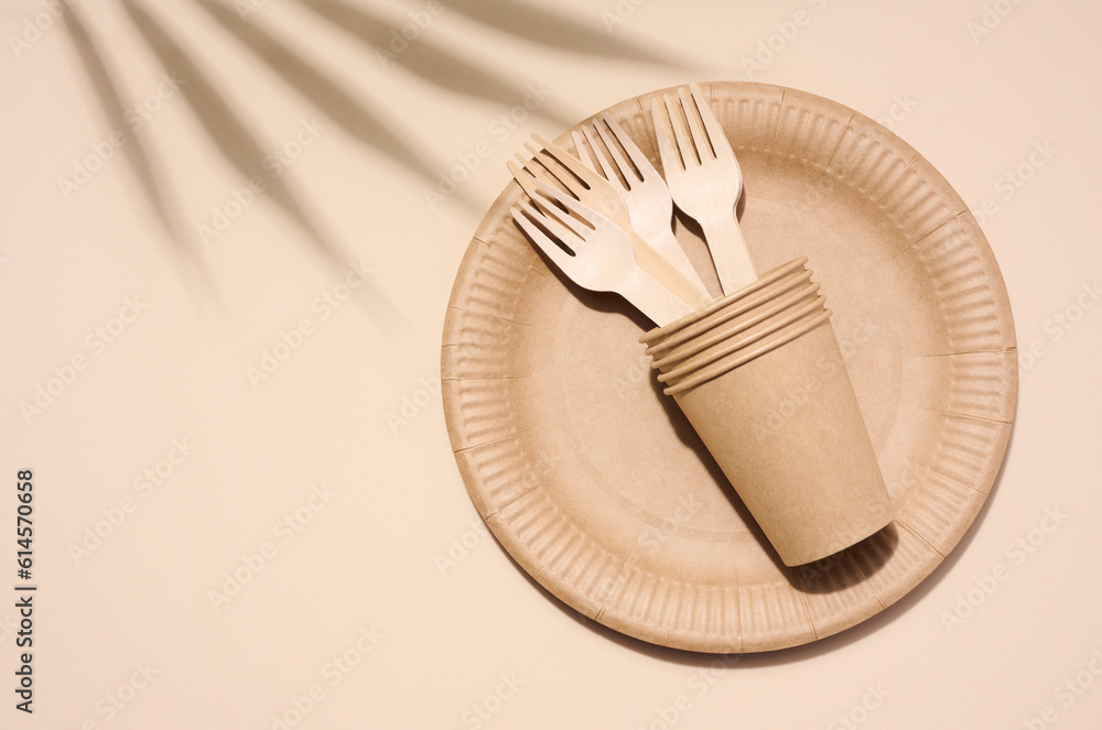 Brown paper plate, stack of glasses and wooden fork, disposable tableware on beige background, top view Brown paper plate, stack of glasses and wooden fork, disposable tableware on beige background, top view