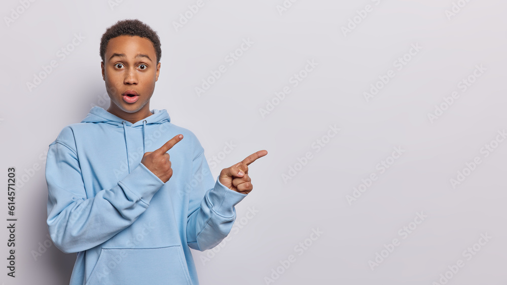 Startled wide eyed striking young man with eyes stands frozen his jaw dropping in sheer horror as gazes at camera points away emphasizing his shock dressed in blue sweatshirt isolated over white wall Startled wide eyed striking young man with eyes stands frozen his jaw dropping in sheer horror as gazes at camera points away emphasizing his shock dressed in blue sweatshirt isolated over white wall