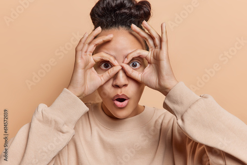 Surprised dark haired Latin woman making circles using her hands around eyes stares impressed has opened mouth wears casual jumper isolated over brown background discovers something amazing. photo
