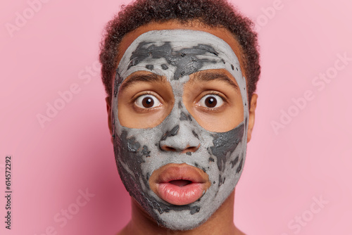 Shocked young man wearing grey clay mask showcases beauty products and cosmetics while serving as model for cosmetologist against pink studio backdrop engaged in his skincare routine poses indoor.