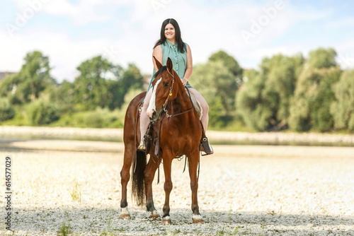 A young woman riding her bay brown horse in front of a rural summer landscape; female equestrian in summertime