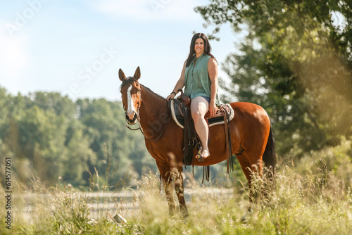 A young woman riding her bay brown horse in front of a rural summer landscape; female equestrian in summertime
