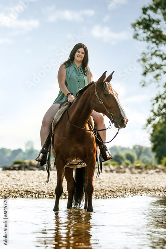 Equestrian and horse cooling down at a hot summer day: A young woman and her bay brown andalusian x arab horse gelding having fun in the water of a river outdoors