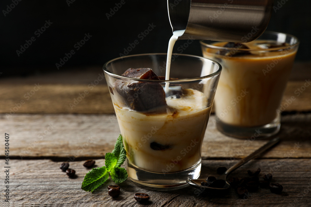 Pouring milk into glass of delicious iced coffee, mint and beans on wooden table Pouring milk into glass of delicious iced coffee, mint and beans on wooden table