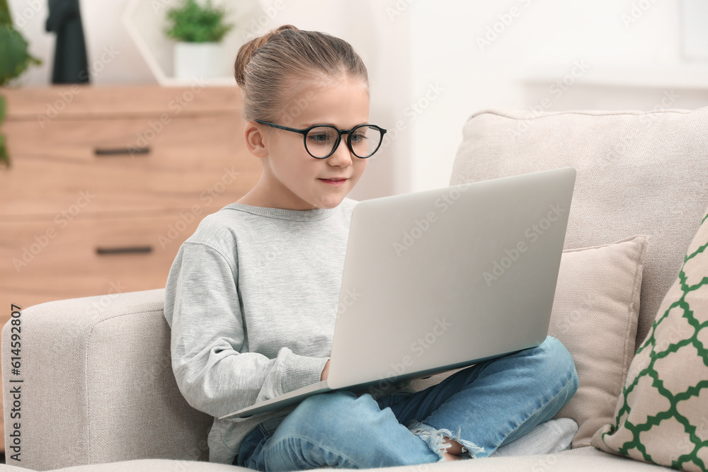 Little girl in glasses using laptop on sofa indoors