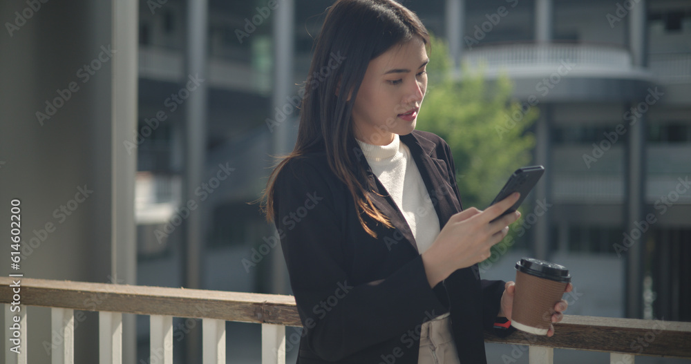 Young Asian business woman in suit holding mobile phone with takeaway coffee
