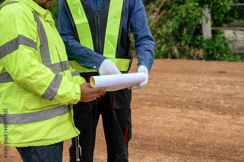 Asian Civil engineers consider and discuss with construction foremen while looking at blueprint papers and schedule targets at highway road construction site.