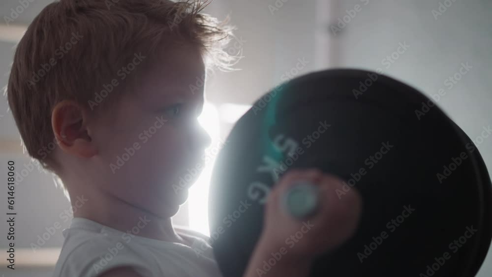 Focused blond toddler pumps arm muscles with heavy barbell standing in gym. Chubby little boy enjoys workout against bright lamp closeup slow motion